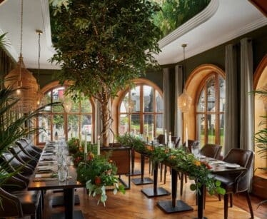Elegant dining room with large arched windows, long wooden tables decorated with greenery, brown leather chairs, hanging lights, and indoor plants, including a tall central tree.