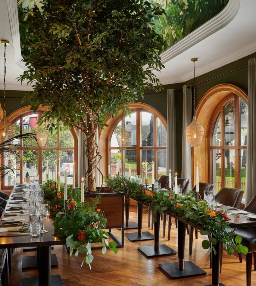 Elegant dining room with large arched windows, long wooden tables decorated with greenery, brown leather chairs, hanging lights, and indoor plants, including a tall central tree.