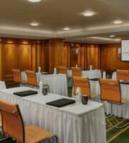 A conference room with rows of tables and chairs, each table set with water bottles, glasses, and folders; a projector screen is visible at the front of the room.