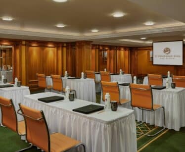 A conference room with rows of tables and chairs, each table set with water bottles, glasses, and folders; a projector screen is visible at the front of the room.