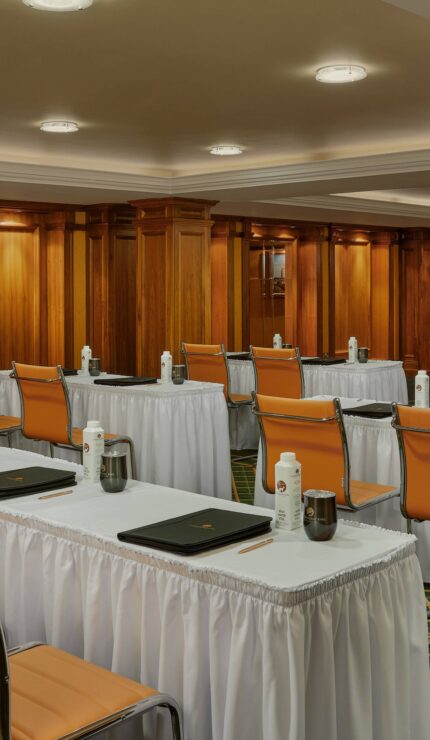 A conference room with rows of tables and chairs, each table set with water bottles, glasses, and folders; a projector screen is visible at the front of the room.