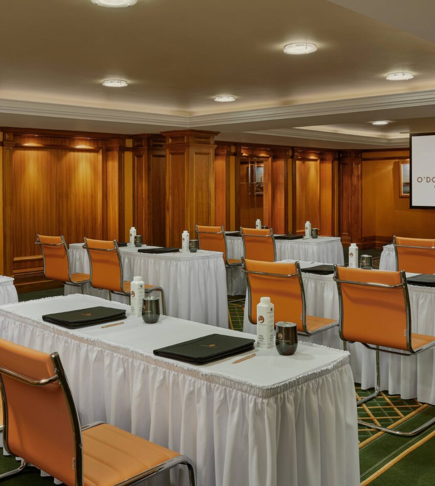 A conference room with rows of tables and chairs, each table set with water bottles, glasses, and folders; a projector screen is visible at the front of the room.