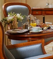 A wooden table set for breakfast with coffee, juice, pastries, and flowers, flanked by upholstered chairs in a well-lit room.