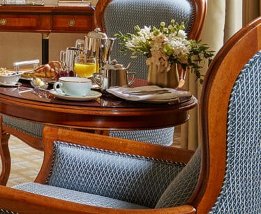 A wooden table set for breakfast with pastries, coffee, orange juice, a blue mug, and a vase of white flowers, surrounded by upholstered chairs.