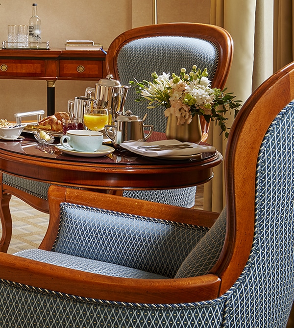 A wooden table set for breakfast with pastries, coffee, orange juice, a blue mug, and a vase of white flowers, surrounded by upholstered chairs.