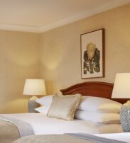 A neatly made bed with white linen, a beige throw cushion, two lamps on bedside tables, and a framed botanical artwork on the wall above the headboard.