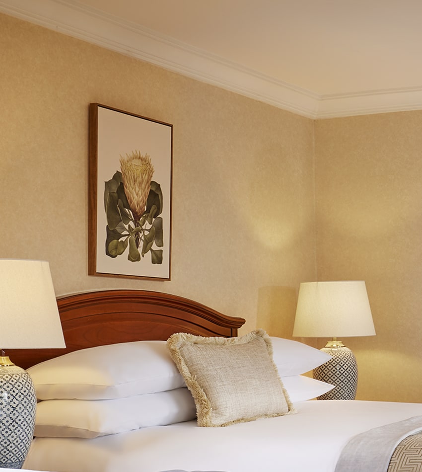 A neatly made bed with a beige pillow, two bedside lamps, and a botanical artwork on the wall above the wooden headboard in a softly lit room.