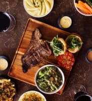 A grilled steak on a wooden board with rocket-topped bread and grilled tomato, surrounded by sides of mashed potato, vegetables, broccoli, and glasses of red wine.