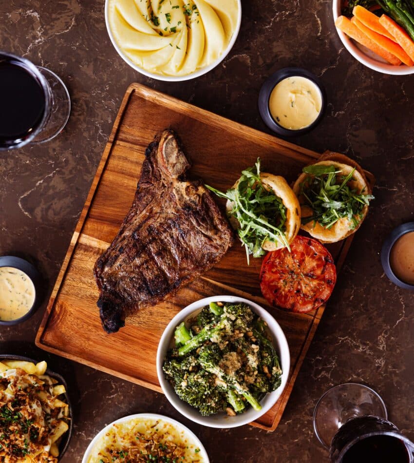 A grilled steak on a wooden board with rocket-topped bread and grilled tomato, surrounded by sides of mashed potato, vegetables, broccoli, and glasses of red wine.