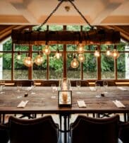 A large wooden dining table is set with glasses and serviettes in a stylish room with leather chairs, hanging lights, and large windows overlooking greenery.