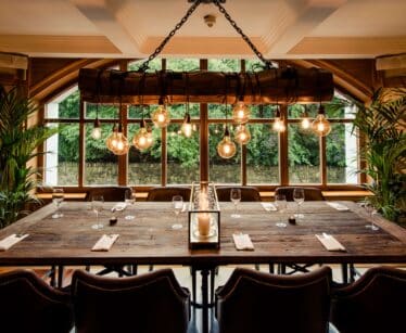 A large wooden dining table is set with glasses and serviettes in a stylish room with leather chairs, hanging lights, and large windows overlooking greenery.