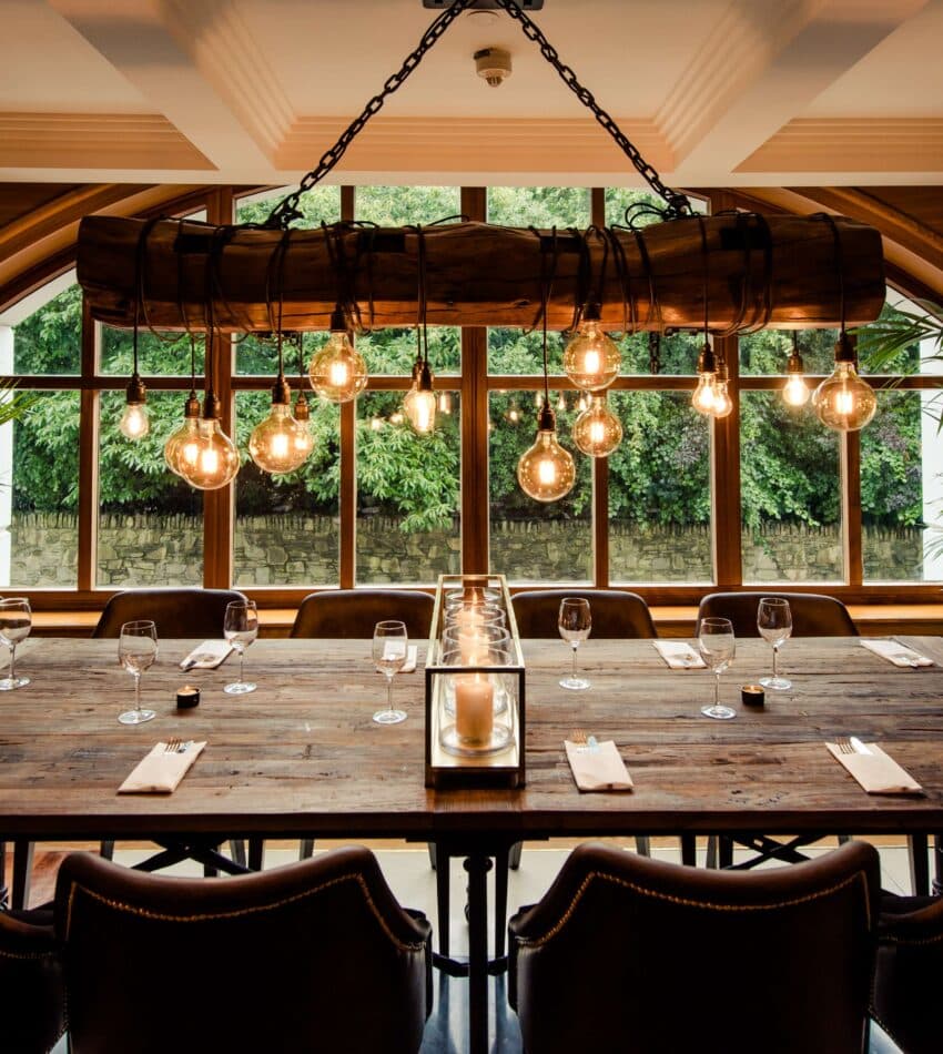 A large wooden dining table is set with glasses and serviettes in a stylish room with leather chairs, hanging lights, and large windows overlooking greenery.