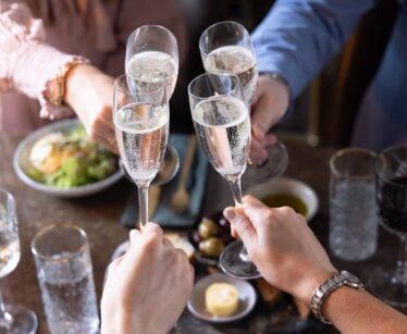 Four people clink glasses of sparkling wine over a table set with food, including a plate with greens, bowls, and glasses of water and red wine.