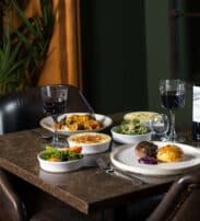 A restaurant table with two place settings, plates of food including meat and vegetables, two glasses of red wine, a bottle of wine, and bowls of assorted side dishes.