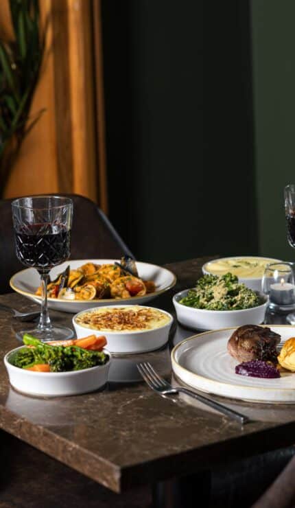 A restaurant table with two place settings, plates of food including meat and vegetables, two glasses of red wine, a bottle of wine, and bowls of assorted side dishes.