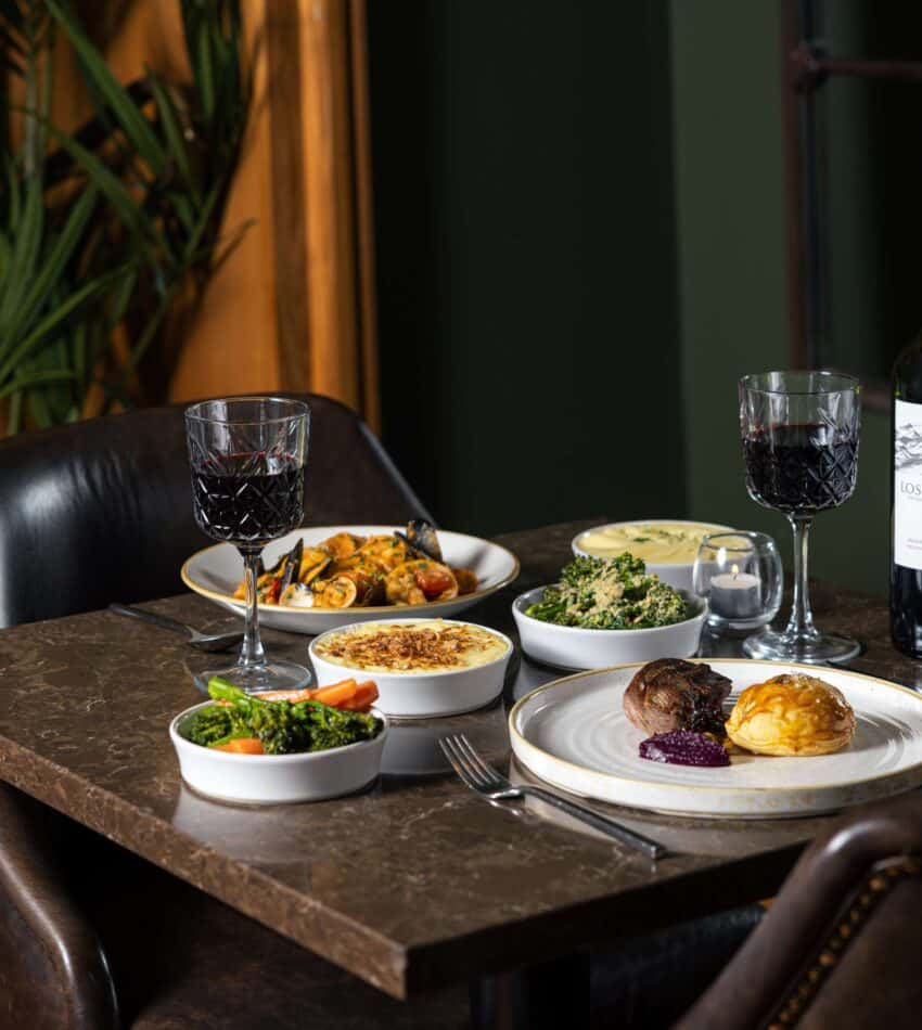 A restaurant table with two place settings, plates of food including meat and vegetables, two glasses of red wine, a bottle of wine, and bowls of assorted side dishes.