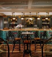 A barman stands behind a fully stocked bar in an upmarket, warmly lit pub with green leather chairs and dark wood furniture.