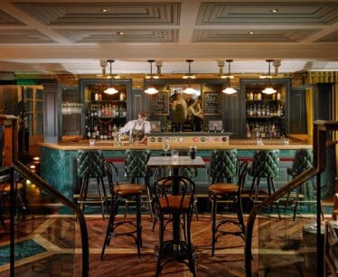 A barman stands behind a fully stocked bar in an upmarket, warmly lit pub with green leather chairs and dark wood furniture.