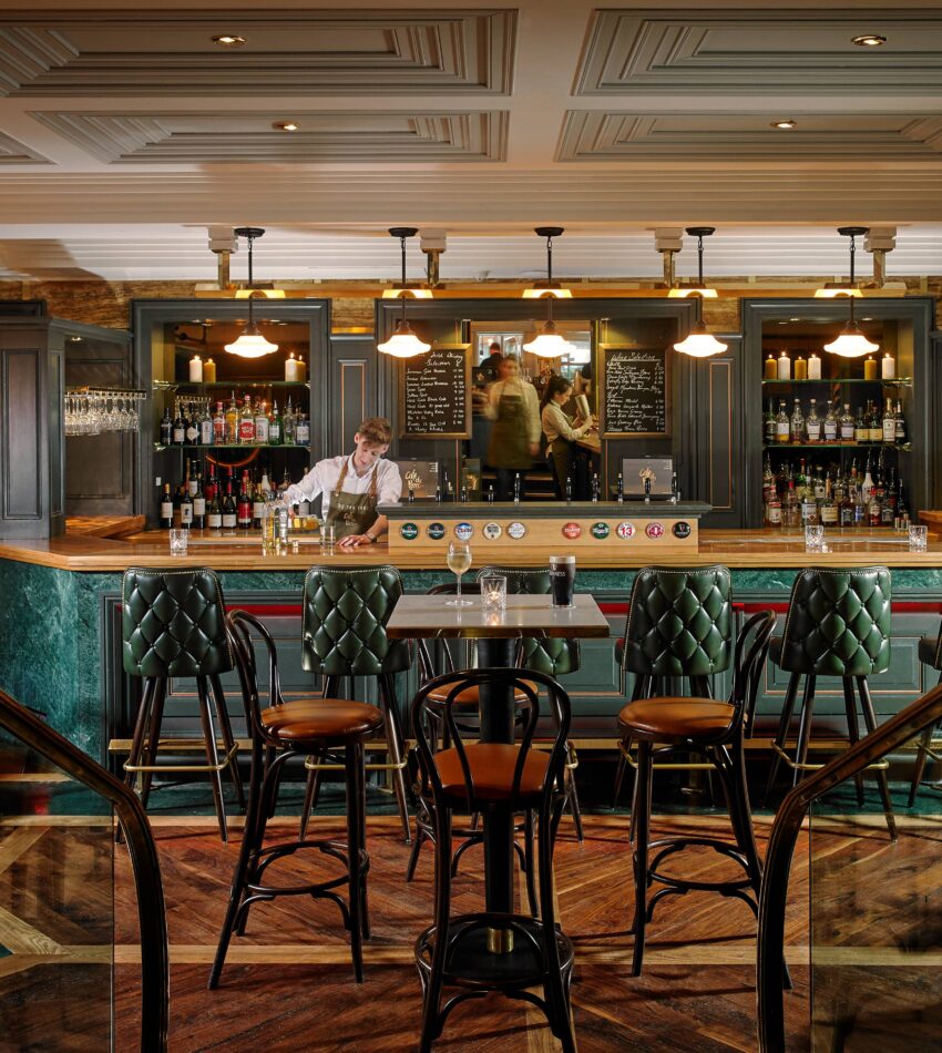 A barman stands behind a fully stocked bar in an upmarket, warmly lit pub with green leather chairs and dark wood furniture.