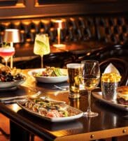 A restaurant table set with sushi, mussels, salad, a burger with chips, assorted drinks including wine, beer, and cocktails, with leather seating in the background.