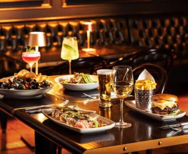 A restaurant table set with sushi, mussels, salad, a burger with chips, assorted drinks including wine, beer, and cocktails, with leather seating in the background.