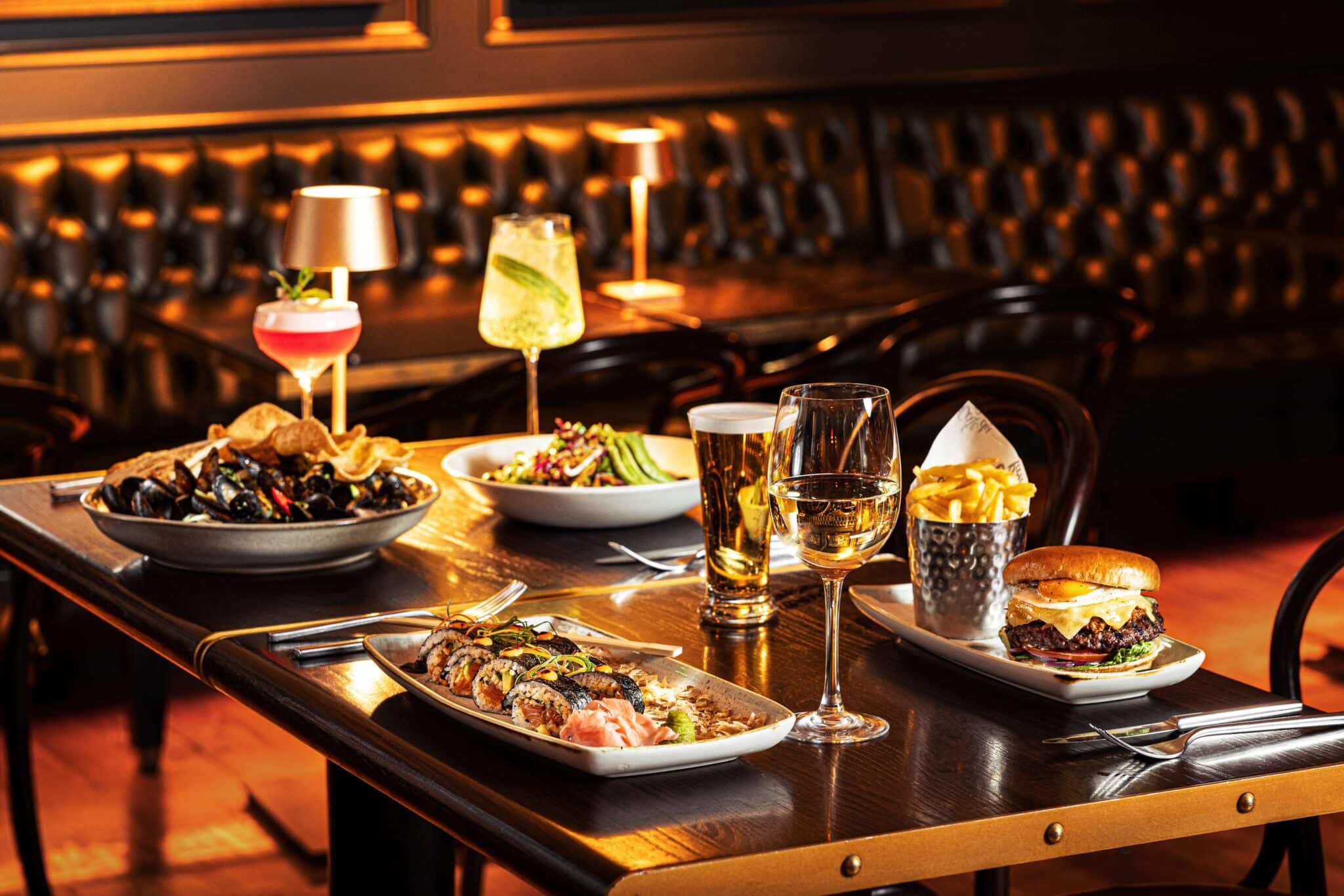 A restaurant table set with sushi, mussels, salad, a burger with chips, assorted drinks including wine, beer, and cocktails, with leather seating in the background.