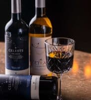 Three wine bottles, two upright and one lying down, are arranged on a marble surface next to a filled cut-glass wine glass.