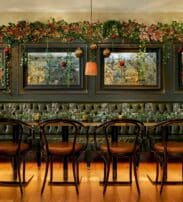 A long dining table with black chairs in front of a dark tufted banquette, decorated with garlands and baubles, set for a meal in an elegant room with mirrors on the wall.