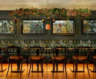 A long dining table with black chairs in front of a dark tufted banquette, decorated with garlands and baubles, set for a meal in an elegant room with mirrors on the wall.