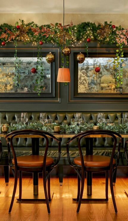 A long dining table with black chairs in front of a dark tufted banquette, decorated with garlands and baubles, set for a meal in an elegant room with mirrors on the wall.