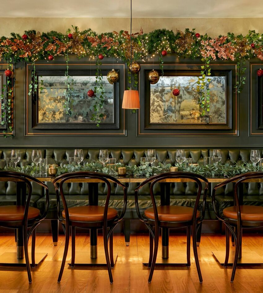 A long dining table with black chairs in front of a dark tufted banquette, decorated with garlands and baubles, set for a meal in an elegant room with mirrors on the wall.