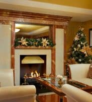 A cosy living room with a lit fireplace, two decorated Christmas trees, cream-coloured sofas, and a glass coffee table, with garland and ornaments adding festive decor.
