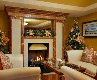 A cosy living room with a lit fireplace, two decorated Christmas trees, cream-coloured sofas, and a glass coffee table, with garland and ornaments adding festive decor.