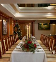 A long dining table set for a formal meal, decorated with candles and a floral centrepiece, in a wood-panelled room with framed pictures and a Christmas tree in the corner.