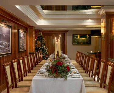 A long dining table set for a formal meal, decorated with candles and a floral centrepiece, in a wood-panelled room with framed pictures and a Christmas tree in the corner.