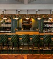A stylish bar with six green cushioned barstools, a wooden countertop, and shelves stocked with bottles and glassware, decorated with greenery above the bar.