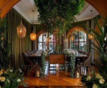 A dining room with a large tree centrepiece, decorative plants, hanging lights, long dining tables set for a meal, and arched windows in the background.