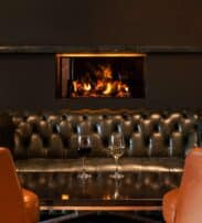 A black leather settee faces a table with two wine glasses; a fireplace with a visible flame is built into the wall behind the settee.