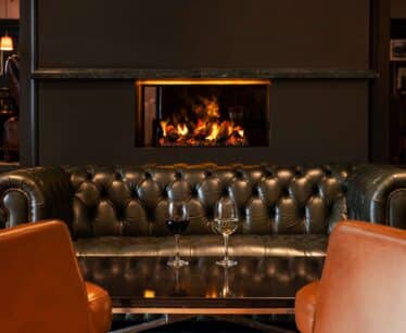 A black leather settee faces a table with two wine glasses; a fireplace with a visible flame is built into the wall behind the settee.