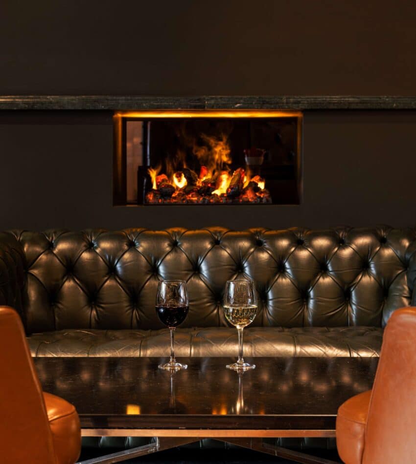 A black leather settee faces a table with two wine glasses; a fireplace with a visible flame is built into the wall behind the settee.