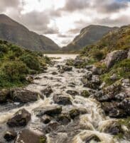 A rocky stream flows through a green valley with mountains in the background under a cloudy sky.