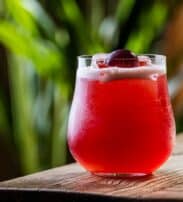 A glass of red cocktail with foam on top and a cherry garnish, placed on a wooden surface with a blurred green background.