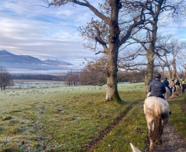 A group of people ride horses along a dirt path in a grassy, tree-lined landscape with mountains and a lake in the background under a cloudy sky.