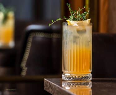 A tall glass of orange cocktail garnished with lemon slices and herbs sits on a marble table, with another blurred drink in the background.
