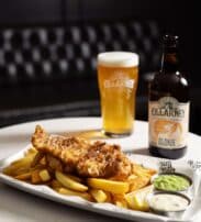 A plate of fish and chips with mushy peas and tartare sauce, served with a glass and bottle of Killarney beer on a white table.