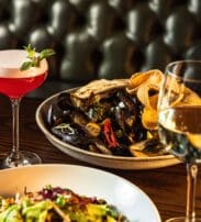 A table with a bowl of mussels and chips, a salad, a pink cocktail garnished with mint, and a glass of white wine, set against a buttoned leather banquette.