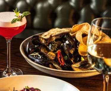 A table with a bowl of mussels and chips, a salad, a pink cocktail garnished with mint, and a glass of white wine, set against a buttoned leather banquette.