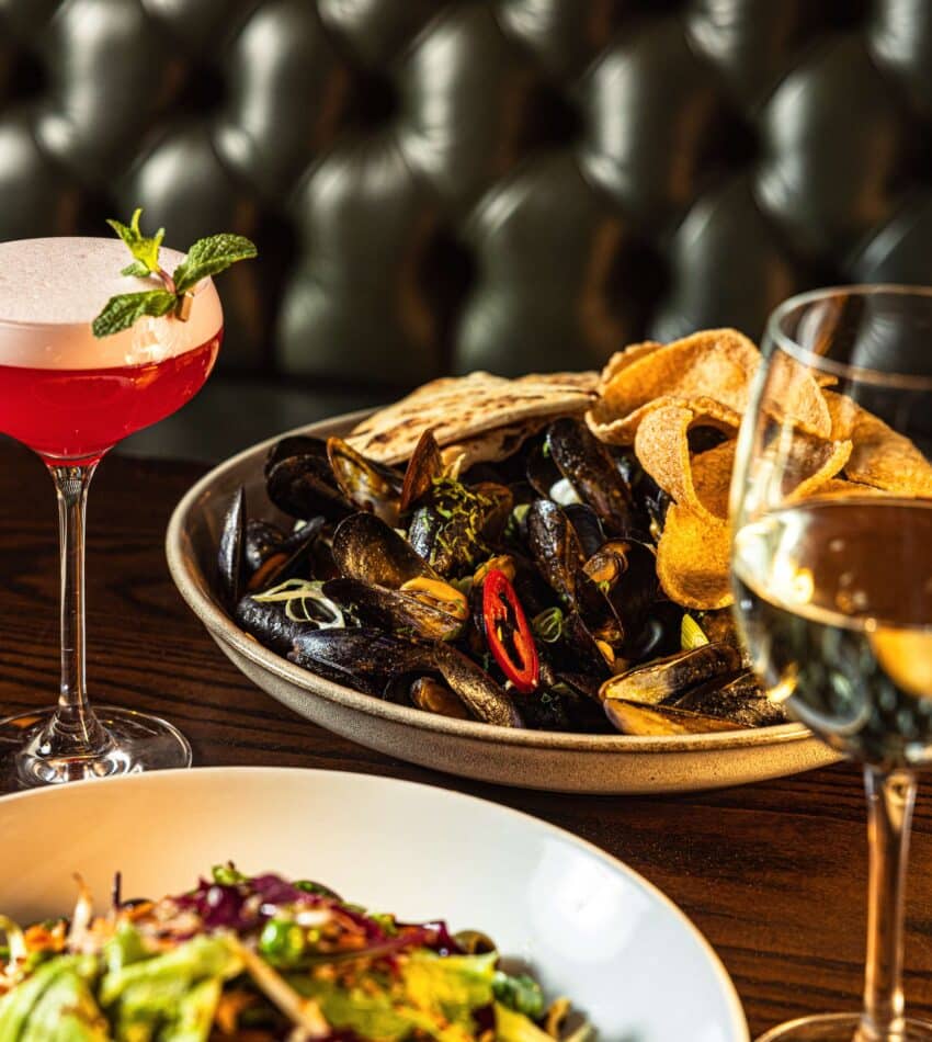 A table with a bowl of mussels and chips, a salad, a pink cocktail garnished with mint, and a glass of white wine, set against a buttoned leather banquette.