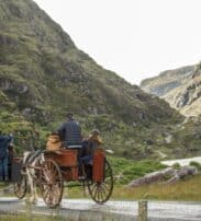 Two horse-drawn carriages with passengers travel along a tarmacked road through a mountainous, green valley landscape.