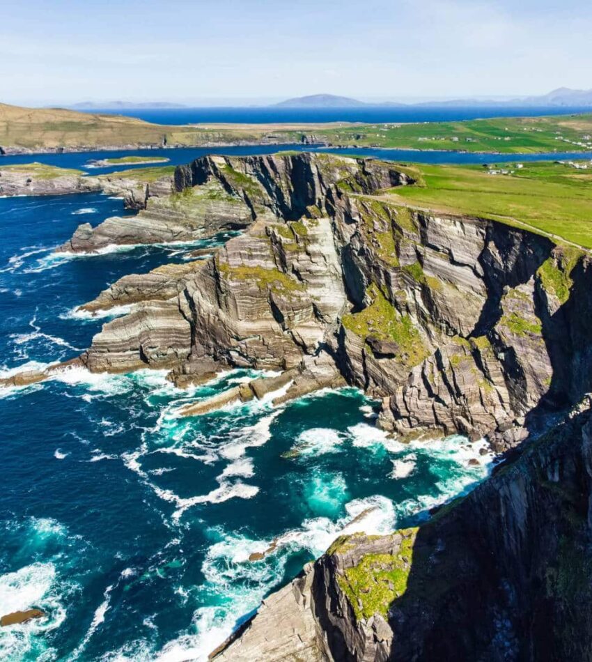 Steep, rocky cliffs line the coastline with waves crashing below, green fields above, and the sea stretching into the distance under a clear sky.